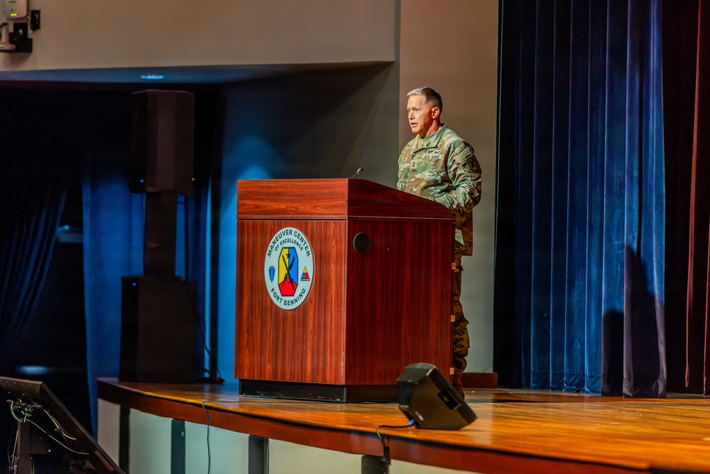 A U.S. Army soldier in OCP (Operational Camouflage Pattern) uniform is speaking at a podium on a stage at Fort Benning's Maneuver Center of Excellence. The wooden lectern prominently displays the Maneuver Center of Excellence insignia. The setting appears to be a formal military auditorium or theater.