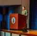 A U.S. Army soldier in OCP (Operational Camouflage Pattern) uniform is speaking at a podium on a stage at Fort Benning's Maneuver Center of Excellence. The wooden lectern prominently displays the Maneuver Center of Excellence insignia. The setting appears to be a formal military auditorium or theater.