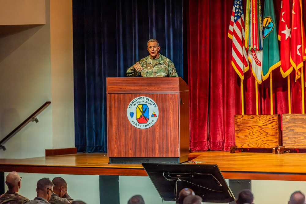 A U.S. Army officer in camouflage uniform is delivering a speech at a podium on a stage at Fort Benning's Maneuver Center of Excellence. The formal military setting includes multiple flags, red and blue curtains, and an audience of uniformed personnel seated in the foreground. The event appears to be an official military ceremony or briefing.