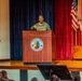 A U.S. Army officer in camouflage uniform is delivering a speech at a podium on a stage at Fort Benning's Maneuver Center of Excellence. The formal military setting includes multiple flags, red and blue curtains, and an audience of uniformed personnel seated in the foreground. The event appears to be an official military ceremony or briefing.