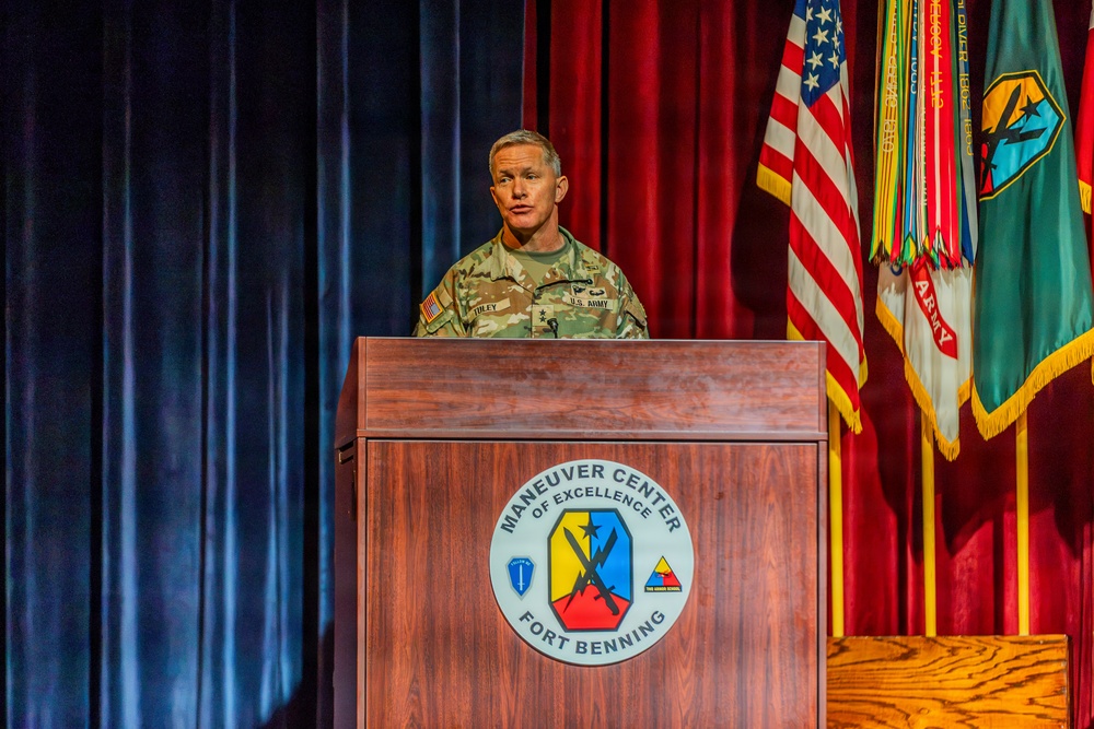 A U.S. Army officer in camouflage uniform is speaking at a podium at Fort Benning's Maneuver Center of Excellence. The speaker appears to hold a general officer rank based on visible insignia on the uniform. The setting is a formal military auditorium with flags and a red curtain backdrop.