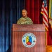 A U.S. Army officer in camouflage uniform is speaking at a podium at Fort Benning's Maneuver Center of Excellence. The speaker appears to hold a general officer rank based on visible insignia on the uniform. The setting is a formal military auditorium with flags and a red curtain backdrop.