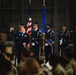 A military ceremony is taking place inside what appears to be a hangar or large indoor facility. An Air Force officer in dress blues is taking a selfie in front of a color guard holding the U.S. flag and an Air Force flag. An audience of uniformed personnel in camouflage uniforms is seated in the foreground.