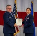 Two United States Air Force officers in dress blue uniforms are jointly holding an official certificate or citation document during what appears to be a formal military awards ceremony. The officer on the right is receiving a medal, visible pinned to their uniform, along with the accompanying citation. The ceremony is taking place in front of a large American flag and a blue Air Force flag.