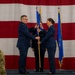 A military change of command ceremony is taking place in front of a large American flag backdrop. Two U.S. Air Force officers in service dress uniforms are exchanging a unit guidon (flag), which is the central ritual of a change of command ceremony. Additional military personnel are present as witnesses, including one officer in Army combat uniform visible in the foreground.