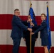 A military change of command ceremony is taking place in front of a large American flag backdrop. Two U.S. Air Force officers in service dress uniforms are exchanging a unit guidon (flag on a staff), which is the traditional centerpiece of a change of command ceremony. A third officer stands in the background holding another flag, while additional personnel are visible in the foreground.
