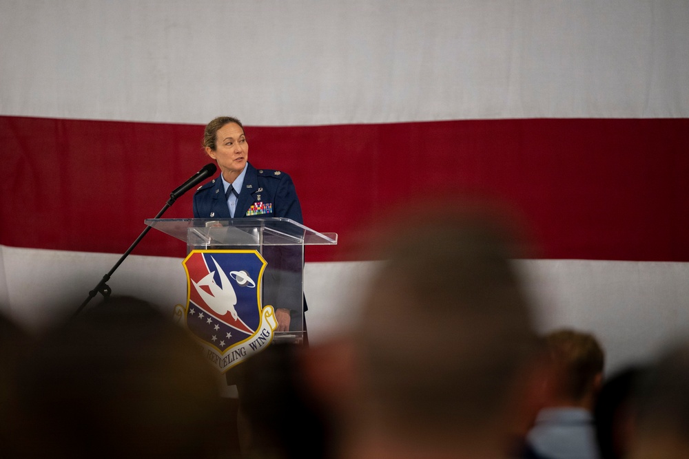 A female U.S. Air Force officer in dress blue uniform is speaking at a podium during a formal military ceremony. The clear acrylic podium displays a unit insignia shield with the partial text '...REFUELING WING' visible. A large American flag serves as a backdrop, and an audience is visible in the foreground.