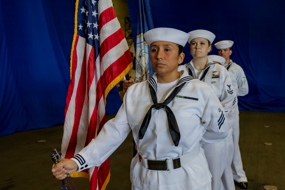 A U.S. Navy color guard is preparing to present or march with the American flag during a formal military ceremony. The lead sailor in the foreground carries the U.S. flag with gold fringe, while three additional sailors in dress white uniforms stand in formation behind her. A blue draped backdrop and a second flag (partially visible) suggest an indoor ceremonial venue.