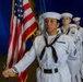 A U.S. Navy color guard is preparing to present or march with the American flag during a formal military ceremony. The lead sailor in the foreground carries the U.S. flag with gold fringe, while three additional sailors in dress white uniforms stand in formation behind her. A blue draped backdrop and a second flag (partially visible) suggest an indoor ceremonial venue.
