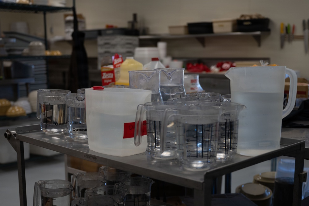 A stainless steel utility cart in what appears to be a commercial kitchen or food preparation area is covered with numerous measuring cups, pitchers, and containers of various sizes. The containers are primarily clear plastic or glass, many with measurement markings visible. The background shows shelving with food products, including a visible 'Zesta' brand package, and additional kitchen equipment.