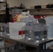 A stainless steel utility cart in what appears to be a commercial kitchen or food preparation area is covered with numerous measuring cups, pitchers, and containers of various sizes. The containers are primarily clear plastic or glass, many with measurement markings visible. The background shows shelving with food products, including a visible 'Zesta' brand package, and additional kitchen equipment.