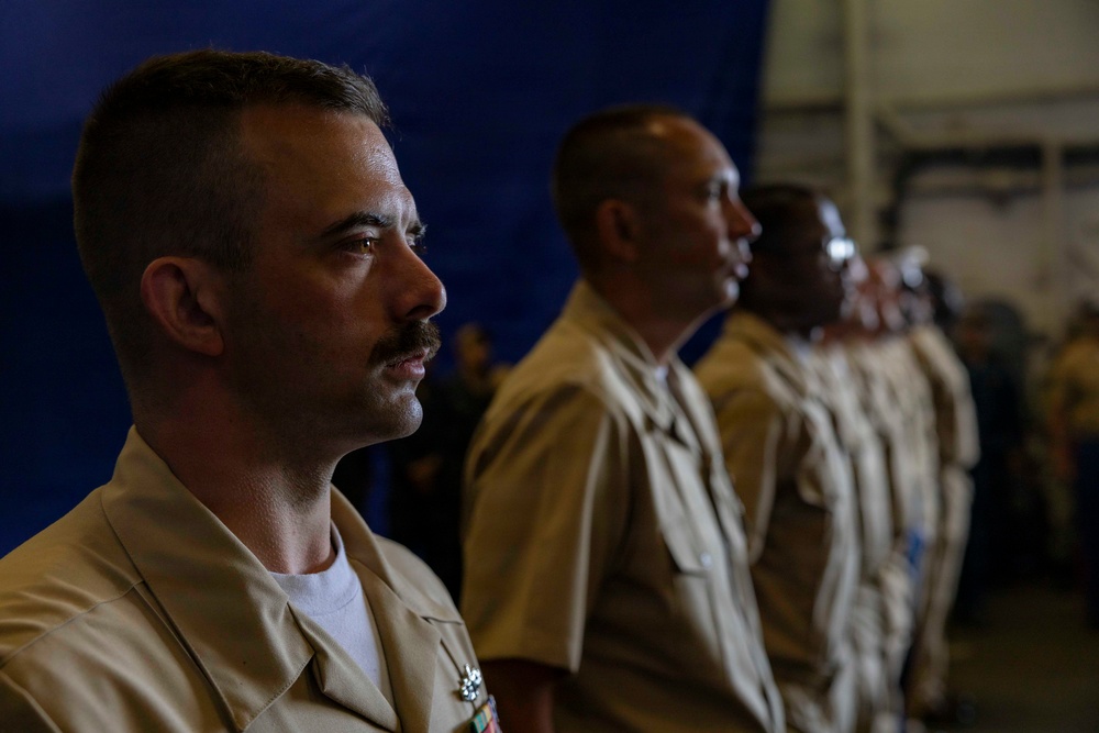 Three military personnel in tan/khaki uniforms are standing in a line, appearing to be at attention during a formal ceremony or event. The foreground subject is a male with a mustache wearing service ribbons on his uniform. The setting appears to be an indoor military facility, possibly a hangar or large bay, with a blue backdrop visible.