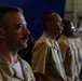 Three military personnel in tan/khaki uniforms are standing in a line, appearing to be at attention during a formal ceremony or event. The foreground subject is a male with a mustache wearing service ribbons on his uniform. The setting appears to be an indoor military facility, possibly a hangar or large bay, with a blue backdrop visible.