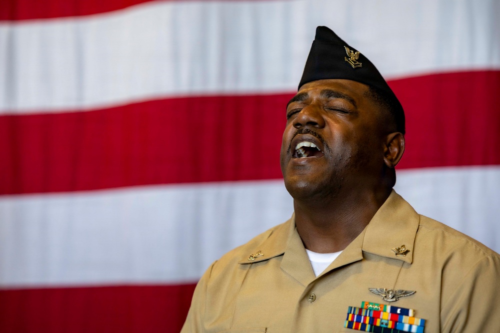 A U.S. Navy enlisted service member in tan/khaki uniform is photographed singing or speaking with eyes closed and mouth open, positioned in front of a large American flag. The individual wears a black garrison cap with an insignia, multiple service ribbons, and aviation-related badges on their uniform. The composition suggests a formal military ceremony or patriotic event.