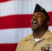 A U.S. Navy enlisted service member in tan/khaki uniform is photographed singing or speaking with eyes closed and mouth open, positioned in front of a large American flag. The individual wears a black garrison cap with an insignia, multiple service ribbons, and aviation-related badges on their uniform. The composition suggests a formal military ceremony or patriotic event.