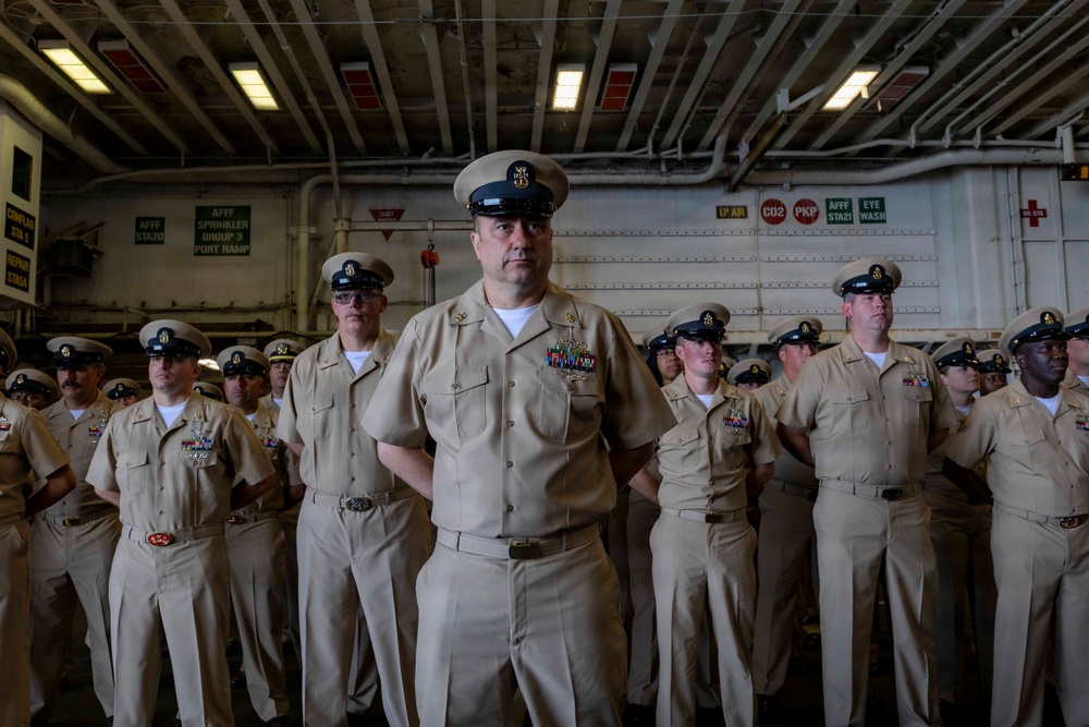 A group of U.S. Navy personnel in khaki service uniforms are standing at attention in what appears to be a ship's hangar bay or interior deck space. The central figure, a senior enlisted sailor with multiple service ribbons and a chief petty officer cover, is prominently positioned in the foreground. The setting includes visible safety signage and overhead piping consistent with a naval vessel interior.