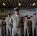 A group of U.S. Navy personnel in khaki service uniforms are standing at attention in what appears to be a ship's hangar bay or interior deck space. The central figure, a senior enlisted sailor with multiple service ribbons and a chief petty officer cover, is prominently positioned in the foreground. The setting includes visible safety signage and overhead piping consistent with a naval vessel interior.