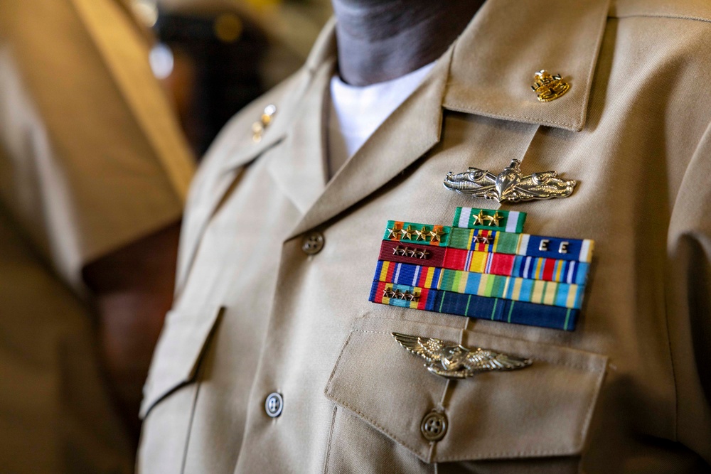 A close-up photograph of a U.S. Navy officer's khaki service uniform showing the chest area with multiple decorations and insignia. The uniform displays a substantial ribbon rack with various awards, two sets of aviation/warfare qualification wings, and collar devices. The image focuses on the decorations and insignia, with the officer's face not visible in the frame.