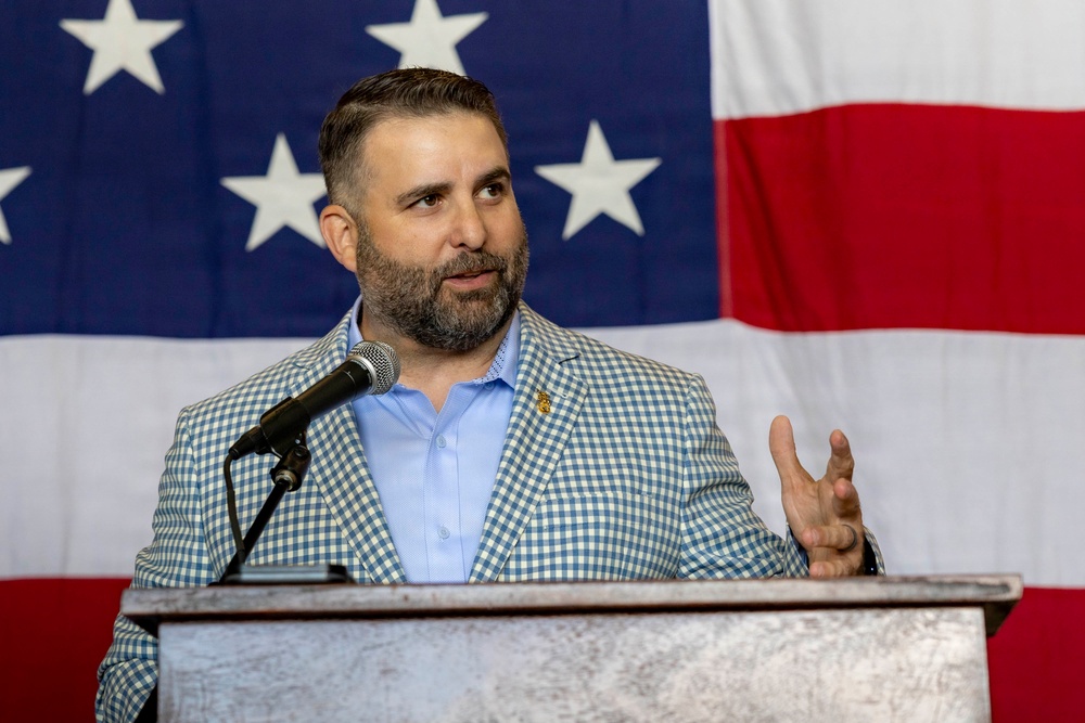 A man with a dark beard and short hair is speaking at a podium with a microphone in front of a large American flag backdrop. He is wearing a blue and white gingham-patterned blazer over a light blue dress shirt, with a small pin on his lapel. His right hand is raised in a gesturing motion, suggesting he is actively delivering a speech or presentation.