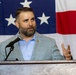 A man with a dark beard and short hair is speaking at a podium with a microphone in front of a large American flag backdrop. He is wearing a blue and white gingham-patterned blazer over a light blue dress shirt, with a small pin on his lapel. His right hand is raised in a gesturing motion, suggesting he is actively delivering a speech or presentation.