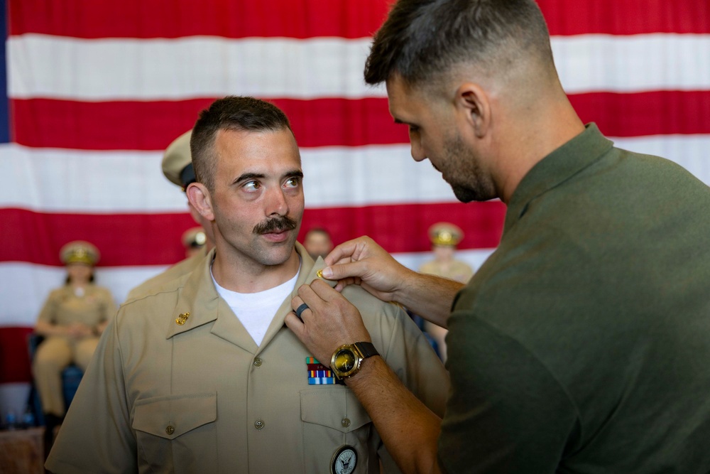 A military pinning ceremony is taking place in front of a large American flag. A person in an olive/green uniform is pinning an insignia or device onto the collar of a service member wearing a tan/khaki Navy uniform with ribbons and medals. The recipient is looking up at the person performing the pinning.