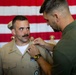 A military pinning ceremony is taking place in front of a large American flag. A person in an olive/green uniform is pinning an insignia or device onto the collar of a service member wearing a tan/khaki Navy uniform with ribbons and medals. The recipient is looking up at the person performing the pinning.