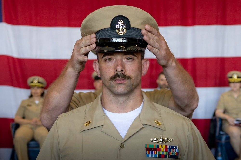 A U.S. Navy Chief Petty Officer is placing his combination cover (cap) on his head during what appears to be a Chief Petty Officer pinning or advancement ceremony. He is wearing the tan/khaki Service Uniform with multiple ribbons, a warfare device, and collar insignia. An American flag is displayed in the background, and other Navy personnel in uniform are seated behind him.
