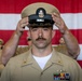 A U.S. Navy Chief Petty Officer is placing his combination cover (cap) on his head during what appears to be a Chief Petty Officer pinning or advancement ceremony. He is wearing the tan/khaki Service Uniform with multiple ribbons, a warfare device, and collar insignia. An American flag is displayed in the background, and other Navy personnel in uniform are seated behind him.