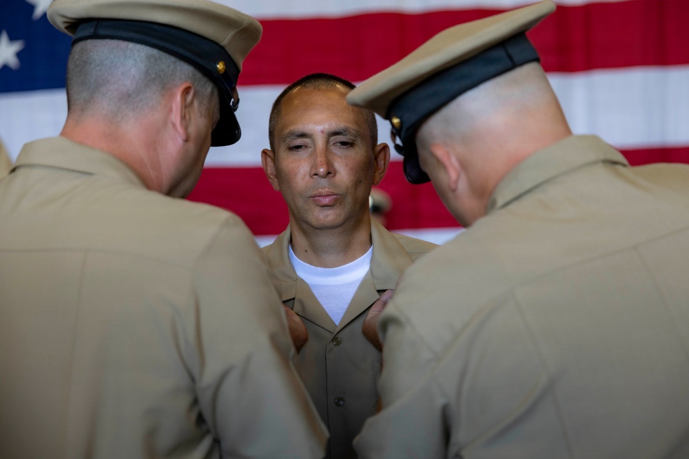 Three military personnel in tan/khaki uniforms are gathered in front of a large American flag. Two officers wearing peaked caps appear to be pinning insignia or decorations onto the uniform of a third service member standing between them. This appears to be a formal military promotion or award ceremony.