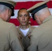 Three military personnel in tan/khaki uniforms are gathered in front of a large American flag. Two officers wearing peaked caps appear to be pinning insignia or decorations onto the uniform of a third service member standing between them. This appears to be a formal military promotion or award ceremony.