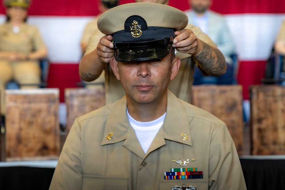 A U.S. Navy Chief Petty Officer is having his combination cover (cap) placed on his head during what appears to be a Chief Petty Officer pinning or advancement ceremony. The subject is wearing the khaki service uniform with visible ribbons, a naval aviator or aircrew warfare device, and collar insignia. The formal ceremony setting includes red and white bunting in the background and wooden chairs.