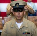 A U.S. Navy Chief Petty Officer is having his combination cover (cap) placed on his head during what appears to be a Chief Petty Officer pinning or advancement ceremony. The subject is wearing the khaki service uniform with visible ribbons, a naval aviator or aircrew warfare device, and collar insignia. The formal ceremony setting includes red and white bunting in the background and wooden chairs.