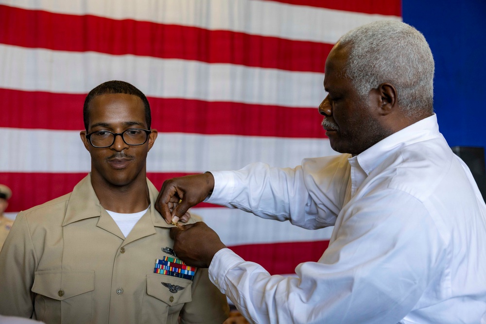 A military promotion or award ceremony is taking place in front of a large American flag backdrop. A civilian man in a white dress shirt is pinning a device or insignia onto the uniform of a U.S. Navy service member wearing a tan/khaki uniform with existing ribbons and wings. The setting appears to be an official military ceremony, likely a promotion, designation, or award pinning.