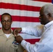 A military promotion or award ceremony is taking place in front of a large American flag backdrop. A civilian man in a white dress shirt is pinning a device or insignia onto the uniform of a U.S. Navy service member wearing a tan/khaki uniform with existing ribbons and wings. The setting appears to be an official military ceremony, likely a promotion, designation, or award pinning.