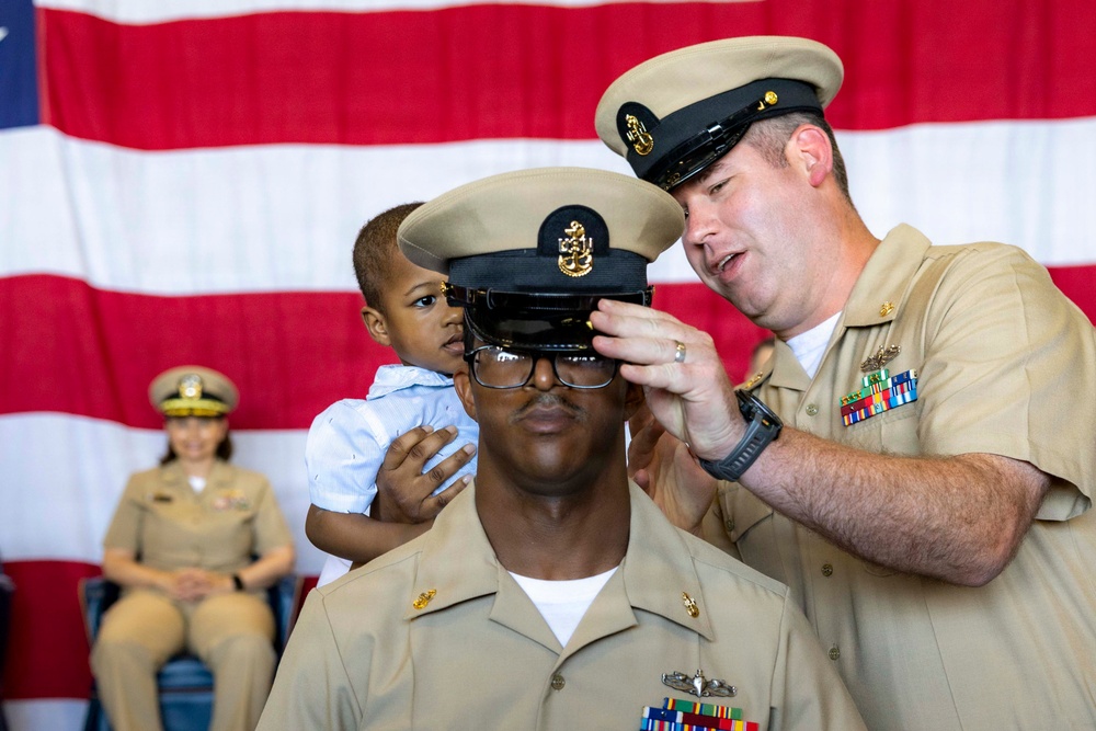 A U.S. Navy promotion ceremony is taking place in front of an American flag backdrop. A senior enlisted sailor is placing a chief petty officer cover (hat) on a newly promoted sailor, while a young child sits on the promotee's shoulders. A female sailor in uniform is seated in the background observing the ceremony.