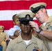 A U.S. Navy promotion ceremony is taking place in front of an American flag backdrop. A senior enlisted sailor is placing a chief petty officer cover (hat) on a newly promoted sailor, while a young child sits on the promotee's shoulders. A female sailor in uniform is seated in the background observing the ceremony.