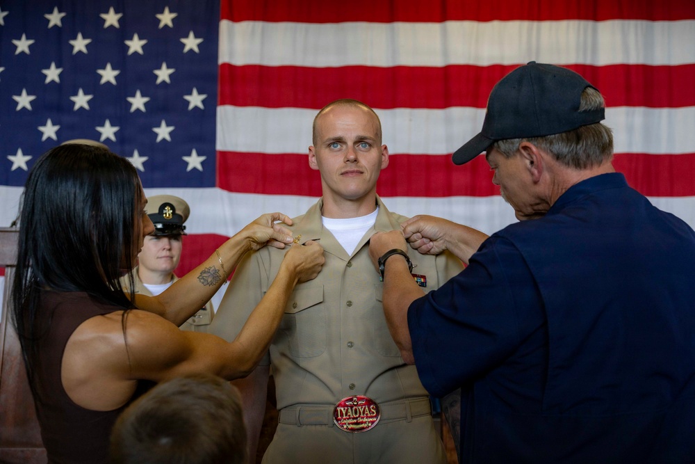 A military promotion or pinning ceremony is taking place in front of a large American flag. A young man in a tan/khaki U.S. Navy uniform is having insignia or rank devices pinned onto his collar by two individuals, likely family members. A uniformed Navy officer stands in the background observing the ceremony.