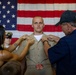 A military promotion or pinning ceremony is taking place in front of a large American flag. A young man in a tan/khaki U.S. Navy uniform is having insignia or rank devices pinned onto his collar by two individuals, likely family members. A uniformed Navy officer stands in the background observing the ceremony.