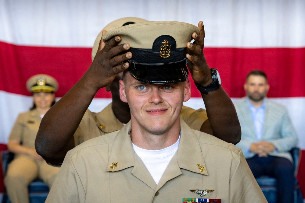 A U.S. Navy Chief Petty Officer pinning/hat ceremony is taking place, where a newly promoted sailor is having the distinctive CPO combination cover (cap) placed on his head by another service member. The subject is wearing a tan/khaki Navy uniform with ribbons, warfare devices, and a name tag. An American flag backdrop and seated audience members in uniform and civilian attire are visible in the background.
