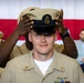 A U.S. Navy Chief Petty Officer pinning/hat ceremony is taking place, where a newly promoted sailor is having the distinctive CPO combination cover (cap) placed on his head by another service member. The subject is wearing a tan/khaki Navy uniform with ribbons, warfare devices, and a name tag. An American flag backdrop and seated audience members in uniform and civilian attire are visible in the background.