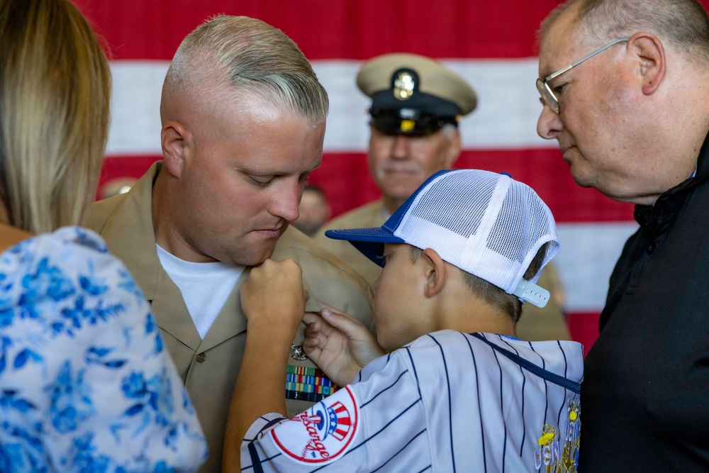 A military promotion or award ceremony is taking place, with a young boy pinning an insignia or device onto a Marine's uniform collar. The Marine is wearing a khaki service uniform with multiple ribbons/medals visible on his chest. An older civilian man assists while a woman in a floral dress and a uniformed Navy officer observe in the background.