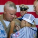 A military promotion or award ceremony is taking place, with a young boy pinning an insignia or device onto a Marine's uniform collar. The Marine is wearing a khaki service uniform with multiple ribbons/medals visible on his chest. An older civilian man assists while a woman in a floral dress and a uniformed Navy officer observe in the background.