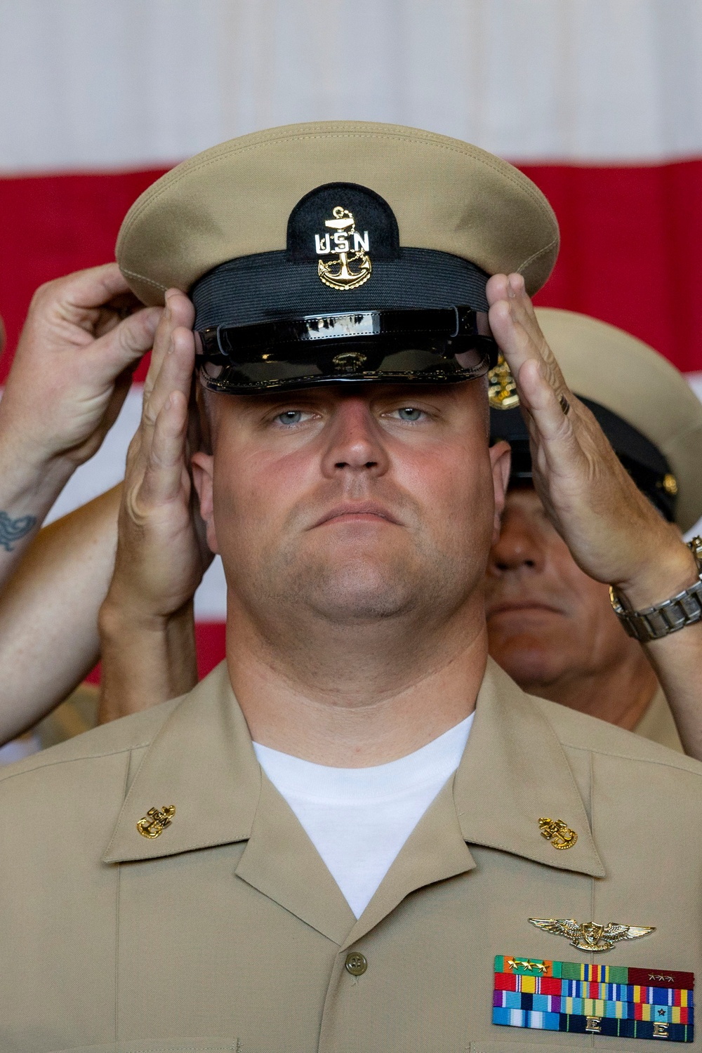 A U.S. Navy Chief Petty Officer is having his combination cover (cap) placed on his head during what appears to be a Chief Petty Officer pinning or frocking ceremony. Two sets of hands from individuals flanking him are simultaneously placing the khaki-topped, black-banded CPO cover onto his head, a traditional part of the CPO initiation ritual. He is wearing the Service Khaki uniform with a Naval Aircrew wings badge and multiple service ribbons.