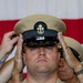 A U.S. Navy Chief Petty Officer is having his combination cover (cap) placed on his head during what appears to be a Chief Petty Officer pinning or frocking ceremony. Two sets of hands from individuals flanking him are simultaneously placing the khaki-topped, black-banded CPO cover onto his head, a traditional part of the CPO initiation ritual. He is wearing the Service Khaki uniform with a Naval Aircrew wings badge and multiple service ribbons.