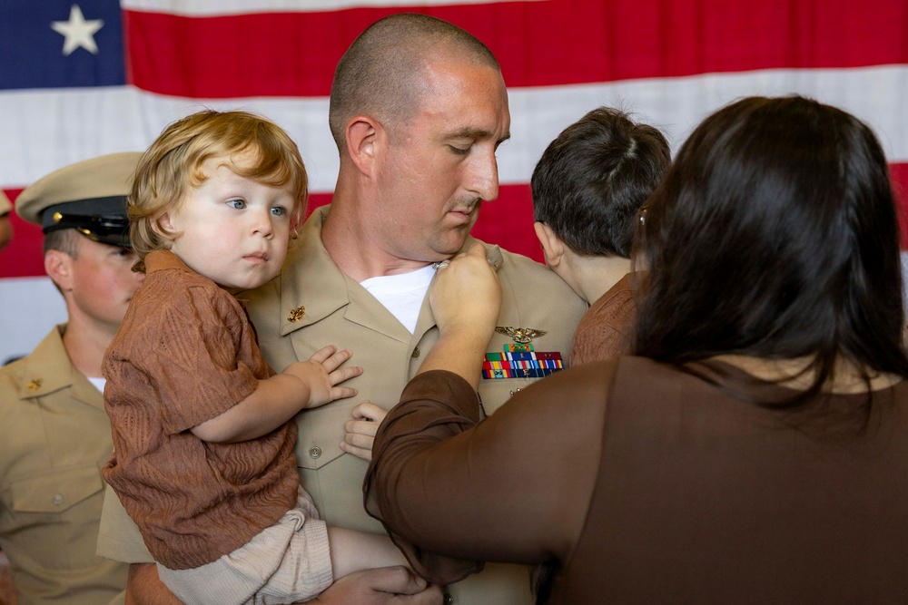 A U.S. Navy officer in tan service uniform is holding a young toddler with curly blonde hair while a woman (facing away) appears to be pinning a new insignia or device onto his uniform. An American flag is displayed prominently in the background, and another uniformed service member is visible to the left. This appears to be a military promotion or pinning ceremony.