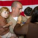 A U.S. Navy officer in tan service uniform is holding a young toddler with curly blonde hair while a woman (facing away) appears to be pinning a new insignia or device onto his uniform. An American flag is displayed prominently in the background, and another uniformed service member is visible to the left. This appears to be a military promotion or pinning ceremony.