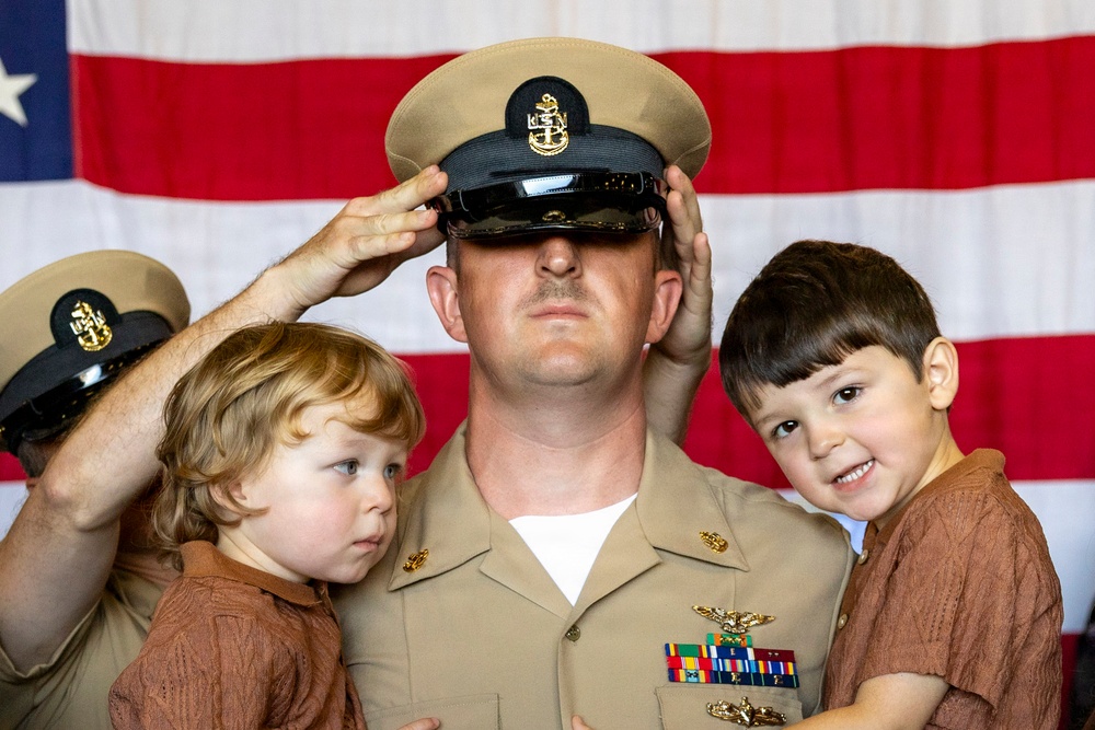 A U.S. Navy Chief Petty Officer is being pinned with his cover (hat) during what appears to be a promotion or advancement ceremony. He is holding two young children, likely his sons, while hands from off-frame place the chief's cover on his head. An American flag serves as the backdrop, indicating a formal military ceremony.