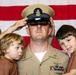A U.S. Navy Chief Petty Officer is being pinned with his cover (hat) during what appears to be a promotion or advancement ceremony. He is holding two young children, likely his sons, while hands from off-frame place the chief's cover on his head. An American flag serves as the backdrop, indicating a formal military ceremony.