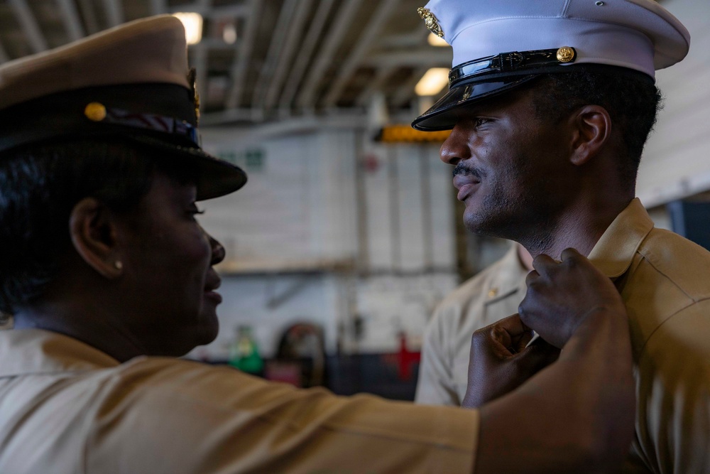 Two U.S. military personnel in khaki/tan uniforms are engaged in what appears to be a pinning or uniform adjustment ceremony. One individual, wearing a cover (hat) with a gold insignia, is adjusting or pinning something onto the collar or chest area of the other, who is wearing a white-topped officer's cover. The setting appears to be aboard a naval vessel, likely in a hangar bay or interior deck space.