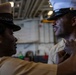 Two U.S. military personnel in khaki/tan uniforms are engaged in what appears to be a pinning or uniform adjustment ceremony. One individual, wearing a cover (hat) with a gold insignia, is adjusting or pinning something onto the collar or chest area of the other, who is wearing a white-topped officer's cover. The setting appears to be aboard a naval vessel, likely in a hangar bay or interior deck space.